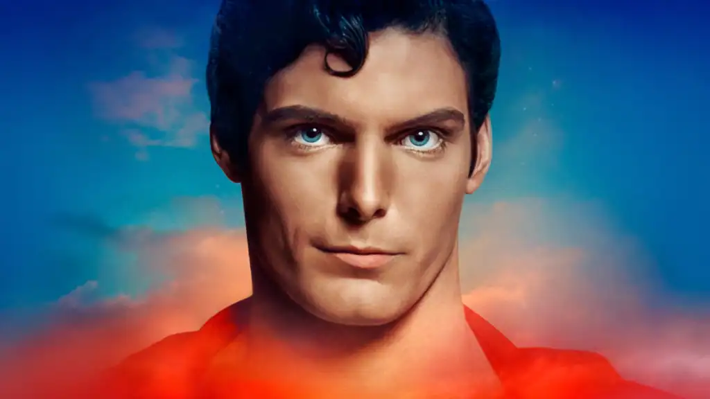 A person with short dark hair and blue eyes stares forward against a blue and orange sky background, wearing a red garment.