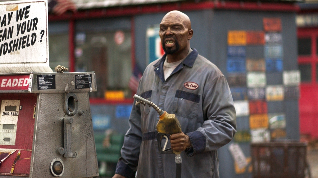 A man in a work uniform holds a gas nozzle, standing next to a gas pump. He appears to be grimacing. The background shows a building with various signs and an American flag.