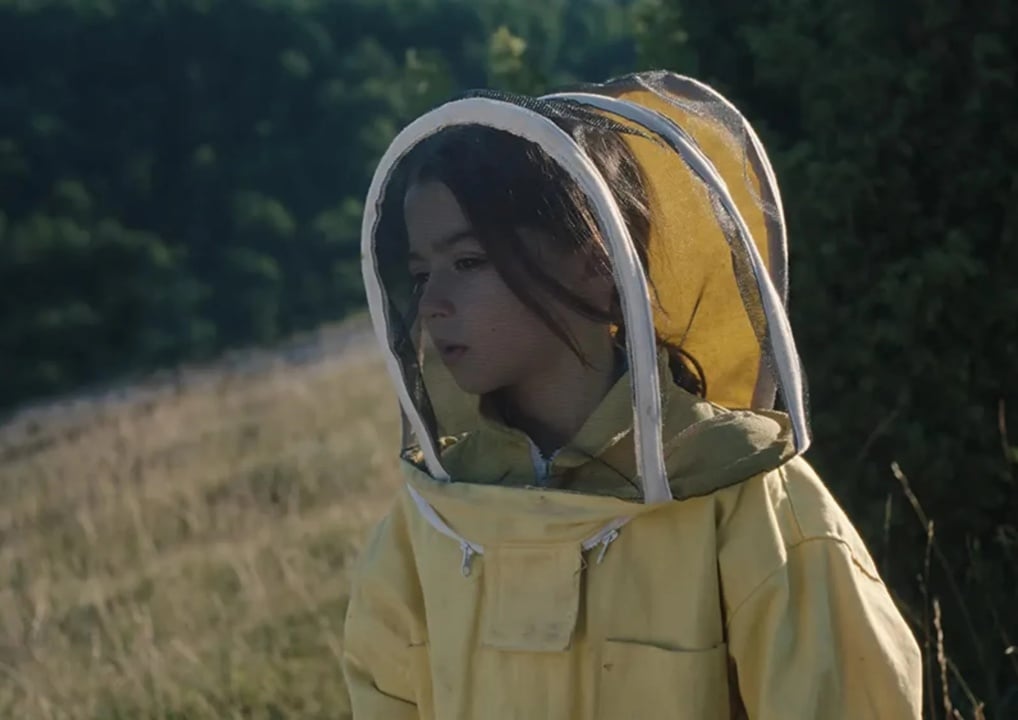 A child wearing a yellow beekeeping suit stands in a grassy field, surrounded by greenery.