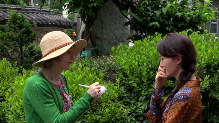 Two women are engaged in conversation outdoors. One is writing on a notepad while the other listens, surrounded by greenery and trees.