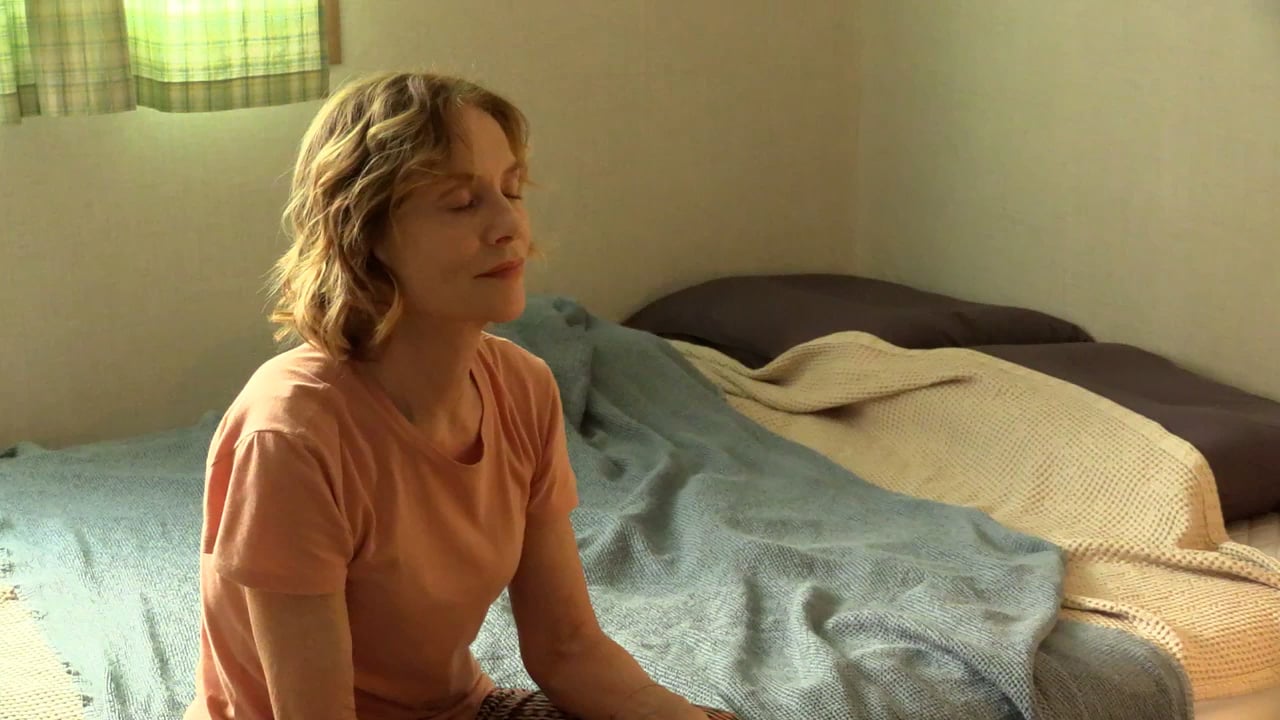 A person sitting on a bed in a softly lit room with eyes closed, wearing a coral shirt. The bed is unmade, covered with a blue blanket and white sheet.