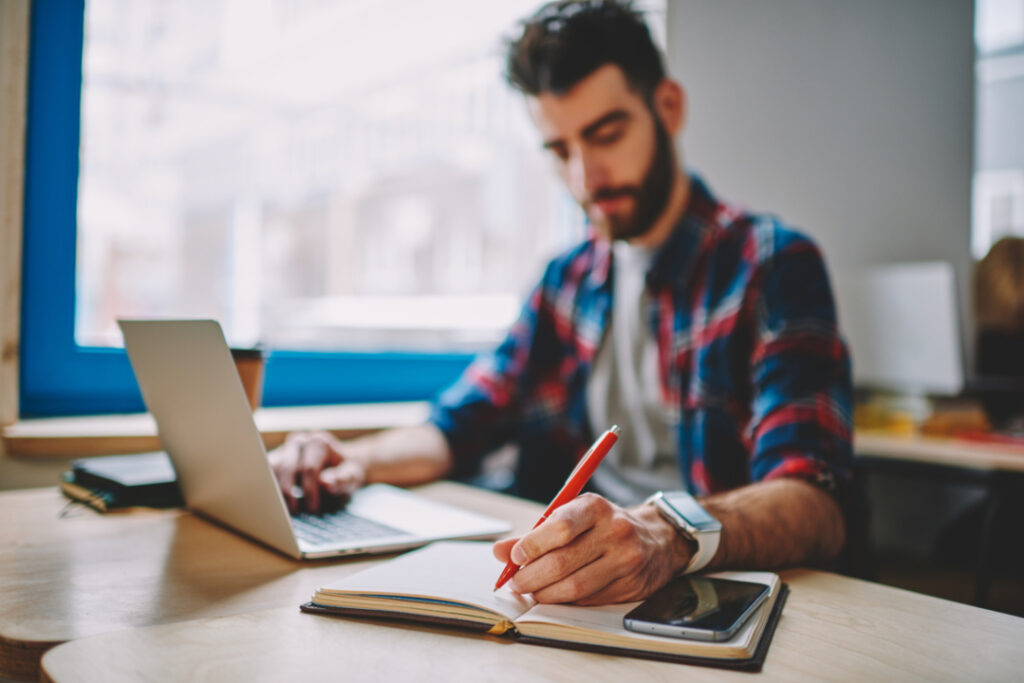 Person in a plaid shirt working on a laptop and writing in a notebook at a desk.