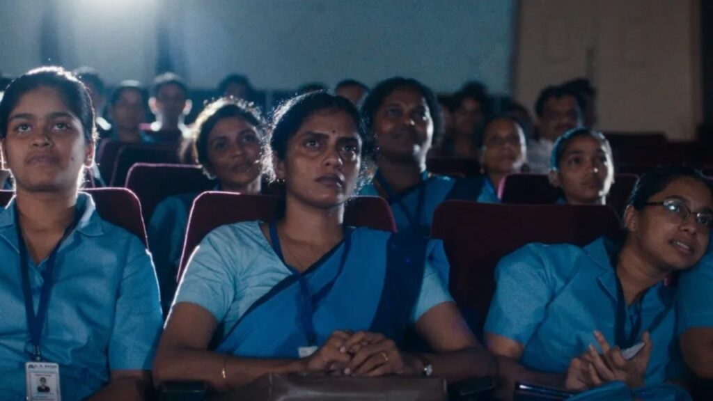 A group of women in blue uniforms sits attentively in a dimly lit auditorium.