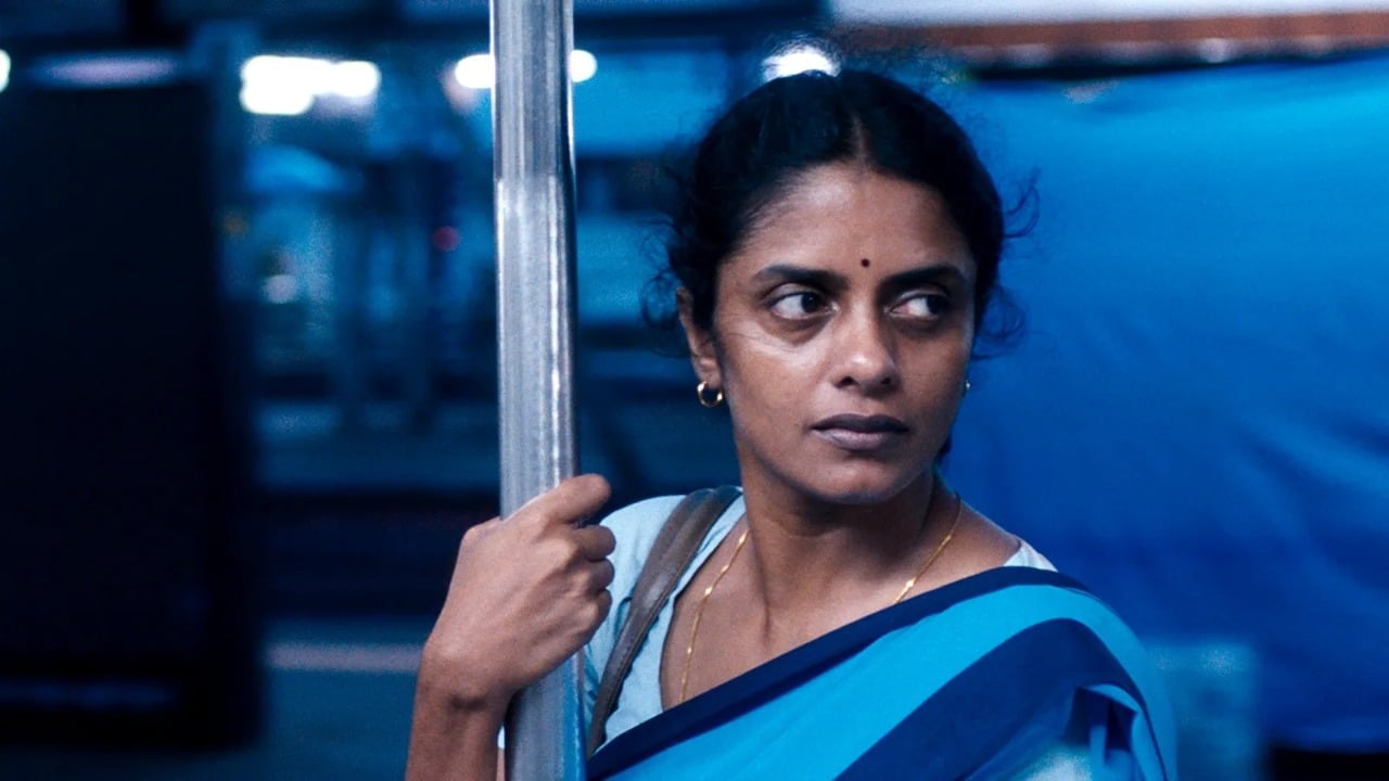 A woman in a blue sari holds a metal pole while looking to the side in a blue-tinged environment.