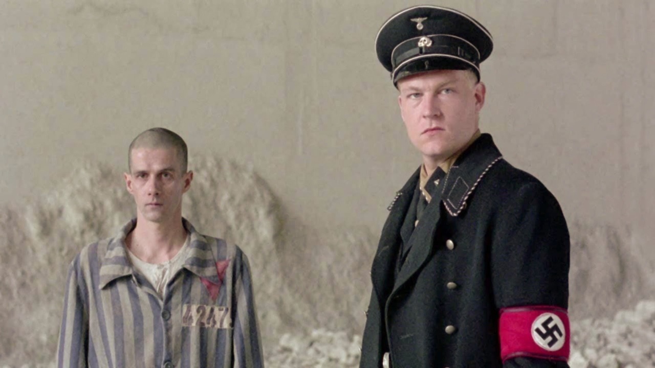 A man in a concentration camp uniform stands next to another in a Nazi uniform with an armband, against a backdrop of concrete walls.