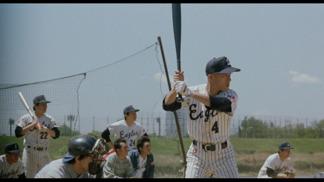 A baseball player in a striped uniform holds a bat, ready to hit. Other players are visible in the background, with a catcher and umpire behind him.