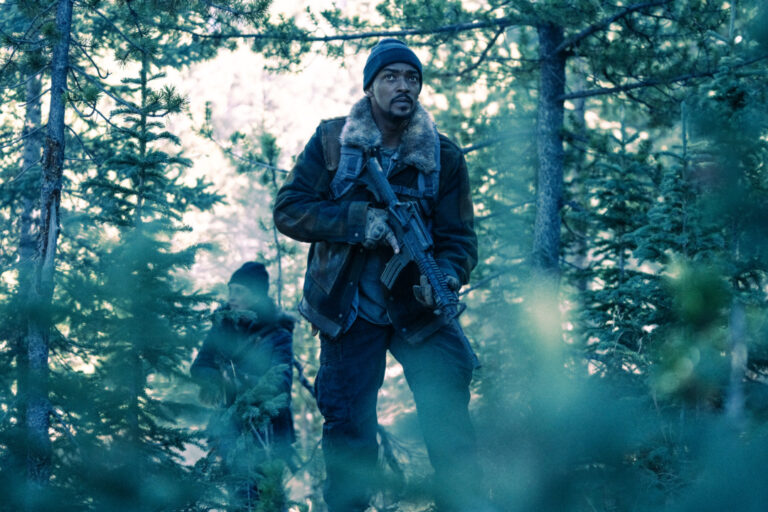 ‘Elevation’ Review: Anthony Mackie Holds The Line In B-Movie Bash Person in a forest wearing a jacket, beanie, and holding a rifle, with another person partially visible in the background.