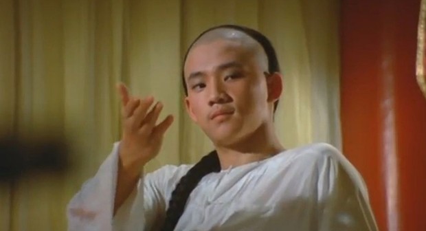 A person in traditional attire with a braid stands indoors, gesturing with one hand.