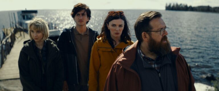 Four people stand on a dock by a lake, with a boat in the background. They are wearing jackets, and the light is dim, suggesting an overcast day.
