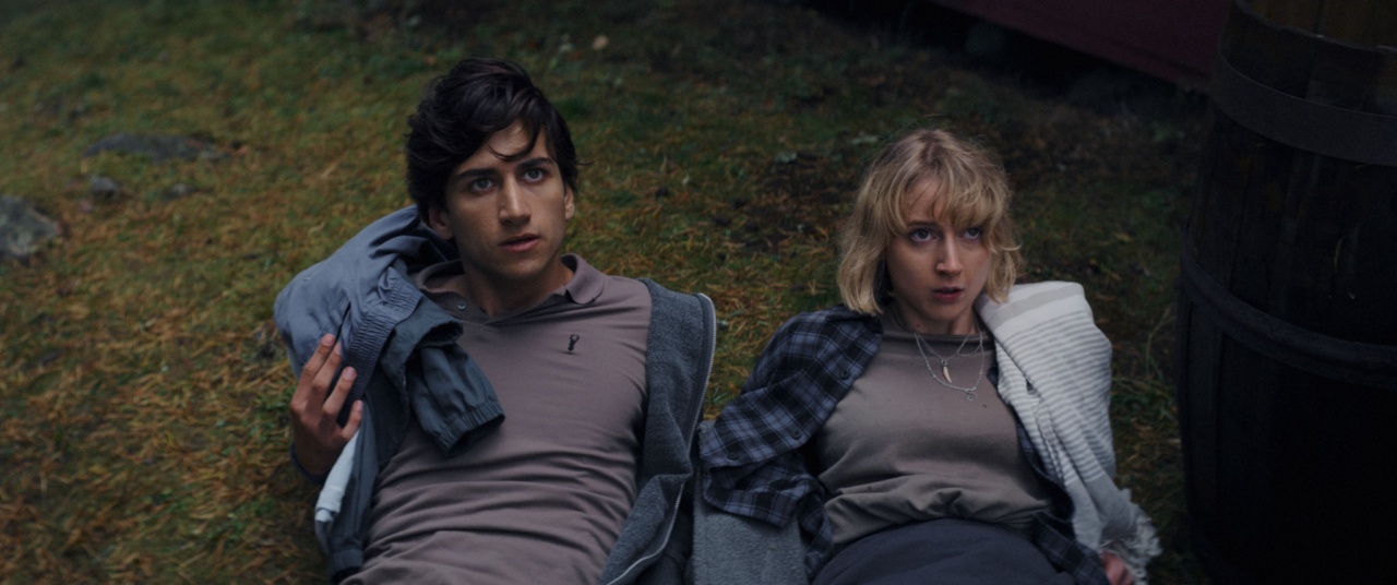 Two people lying on the grass, looking up with surprised expressions. They each wear neutral-colored shirts and have jackets draped over them.