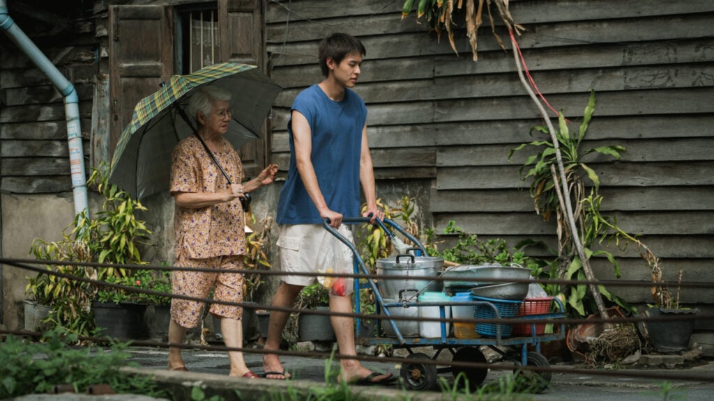 A young man pushes a cart with pots and pans while an elderly woman holds an umbrella, walking behind him in front of a wooden building with plants.