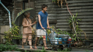 A young man pushes a cart with pots and pans while an elderly woman holds an umbrella, walking behind him in front of a wooden building with plants.