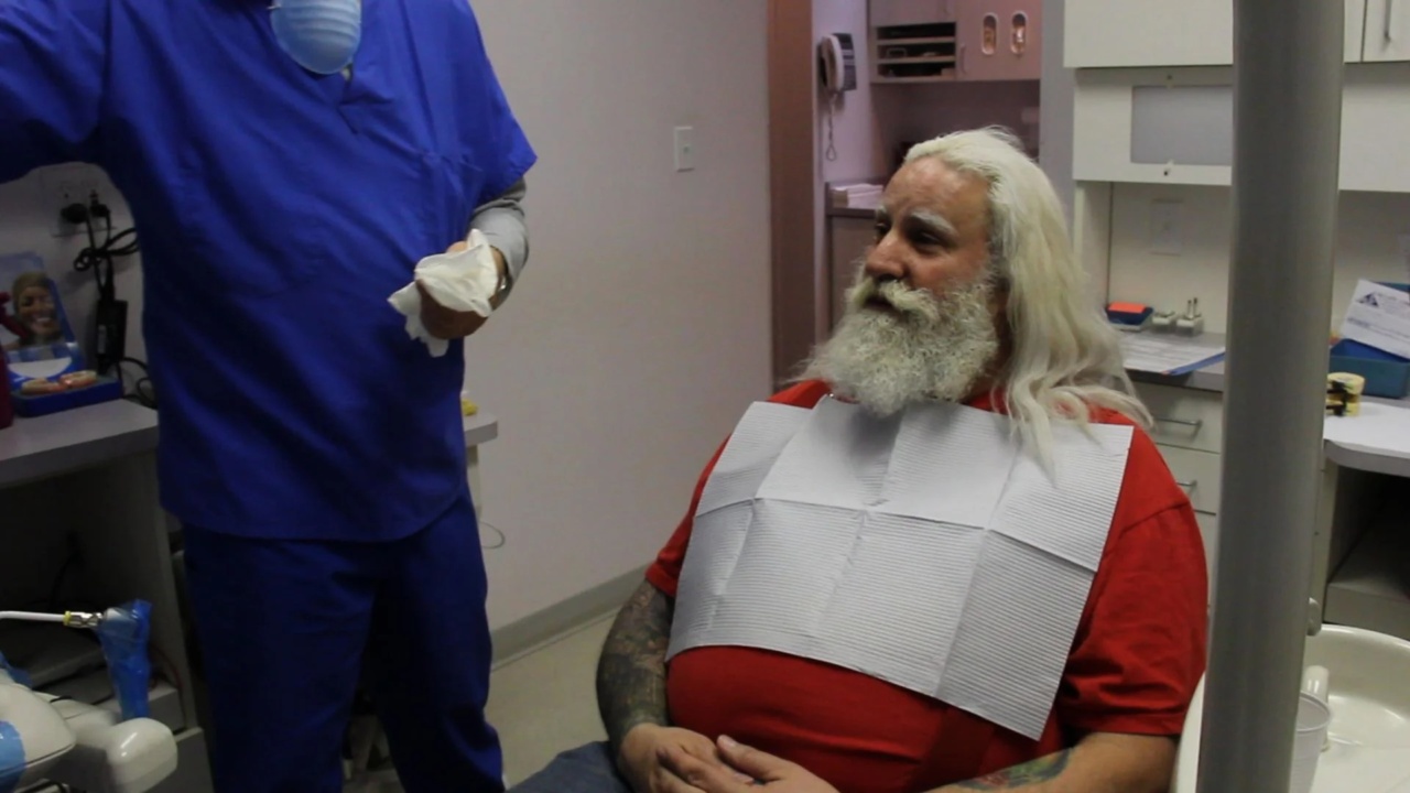 A man with long white hair and beard sits in a dental chair wearing a red shirt and dental bib. A person in blue scrubs stands nearby holding a cloth.