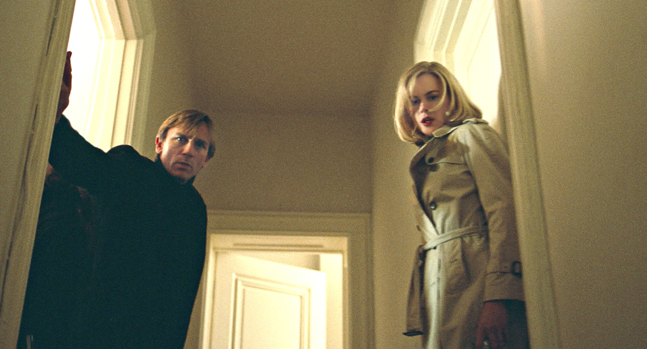 A man and a woman stand at the top of a staircase, looking downward. The woman wears a trench coat, and the man is partially hidden behind a doorway.