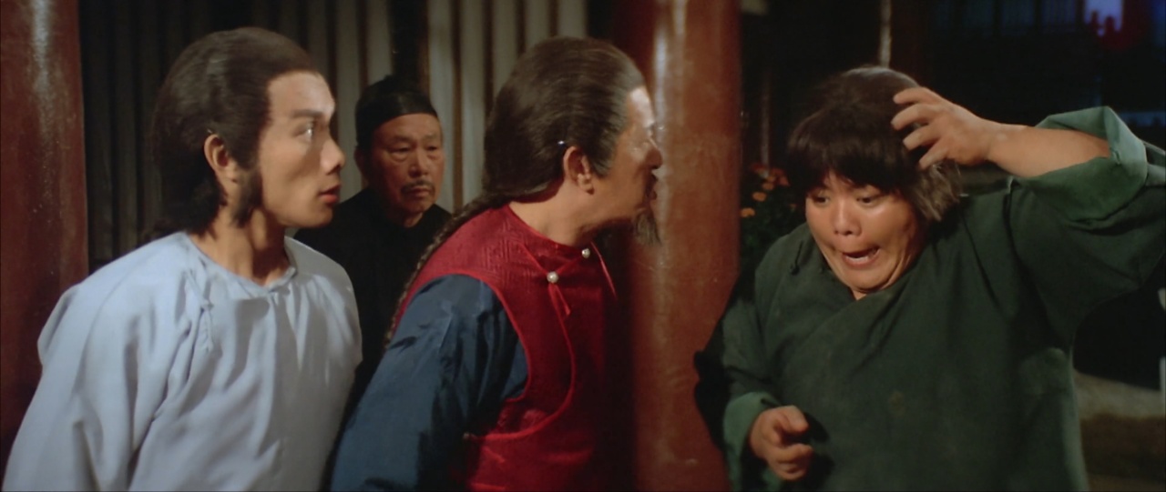 Four people in period costumes stand together indoors, with one person dramatically touching their head.