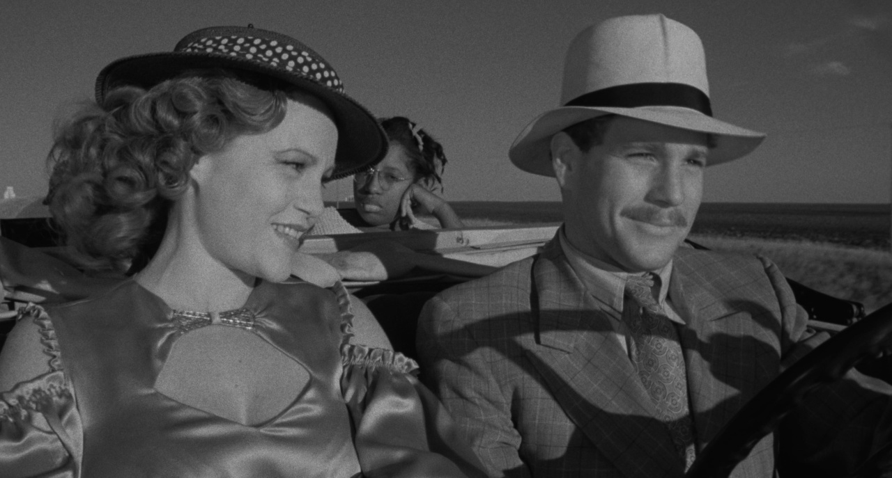 A woman and a man in hats are smiling while sitting in a car. A child in the backseat looks out the window.