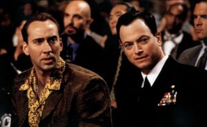 Two men are seated in a crowded auditorium, both wearing jackets. One man has a patterned shirt, while the other wears a decorated suit.