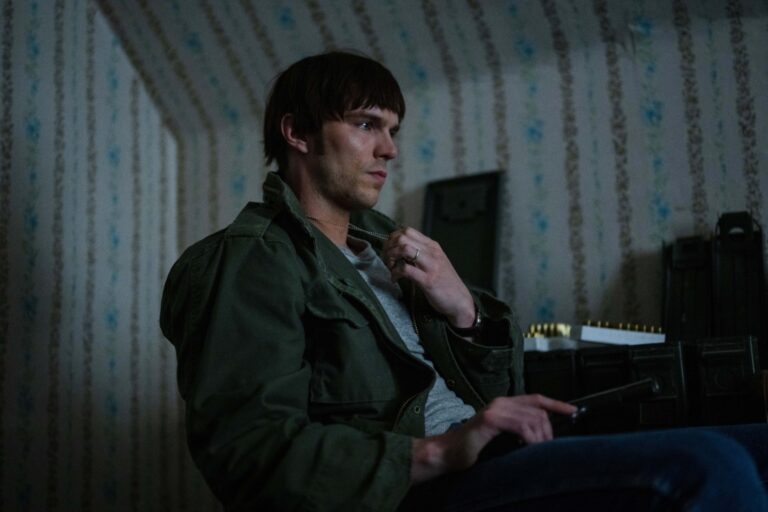 A person in a dimly lit room sits holding a phone, wearing a dark green jacket and a gray shirt, with an open case containing various items nearby.