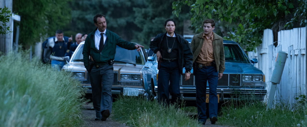 A group of people, including two in law enforcement attire, walk beside parked cars on a grassy path, surrounded by trees and a white fence.