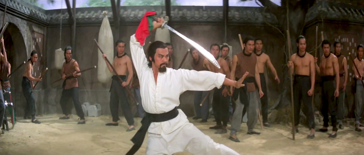 A martial artist in a white uniform wields a sword in front of a group of people in a courtyard setting.