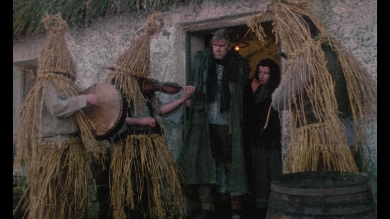 People in straw costumes play music outside a rustic building, while two others watch from the doorway.