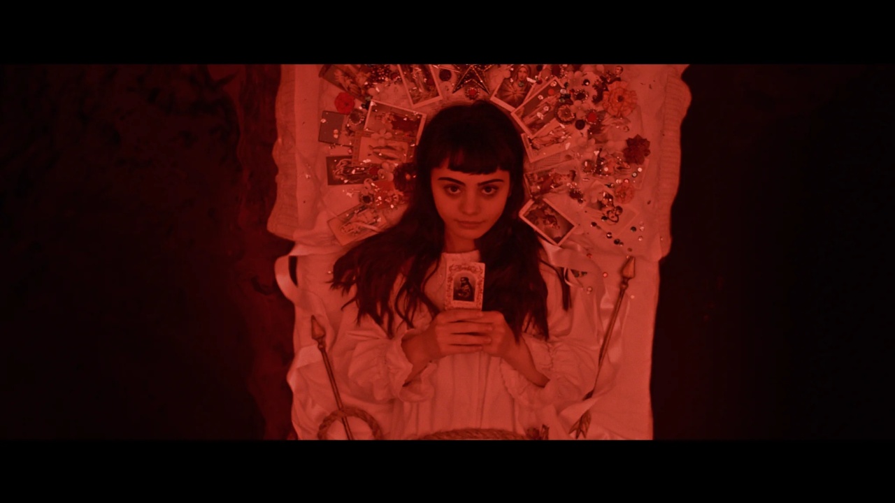 A woman with dark hair, lying in a red-tinted setting, holds a card in her hands. She is surrounded by decorative elements.