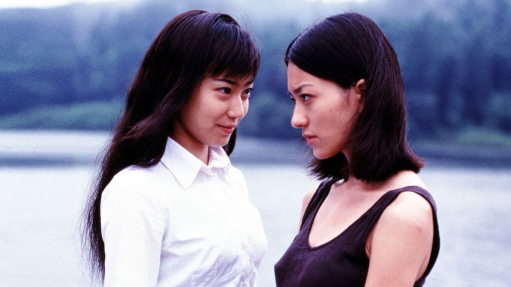 Two women stand face to face by a body of water, one in a white shirt and the other in a black tank top, with trees blurred in the background.