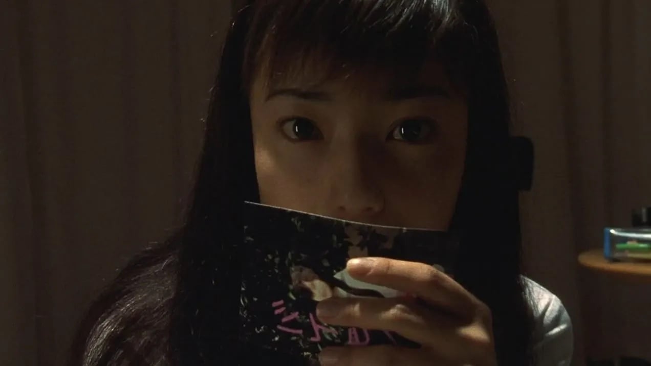 A person with long hair holds up a booklet, partially covering their face, while looking directly at the camera. The background is dimly lit.