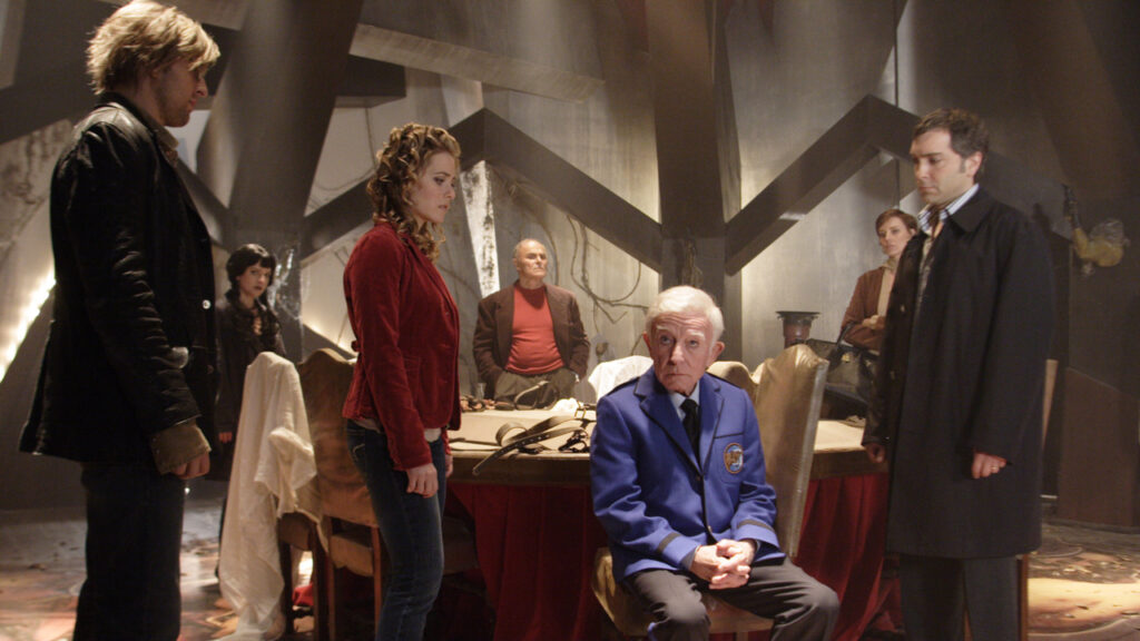 A group of people stand around an older man seated in a room with dim lighting and industrial decor.