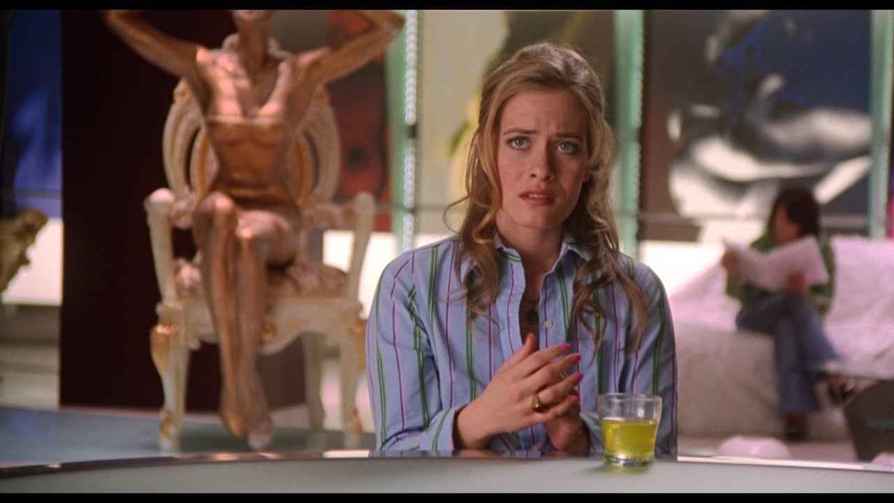 A woman sits at a table with a drink, looking concerned. A statue-like figure is in the background.