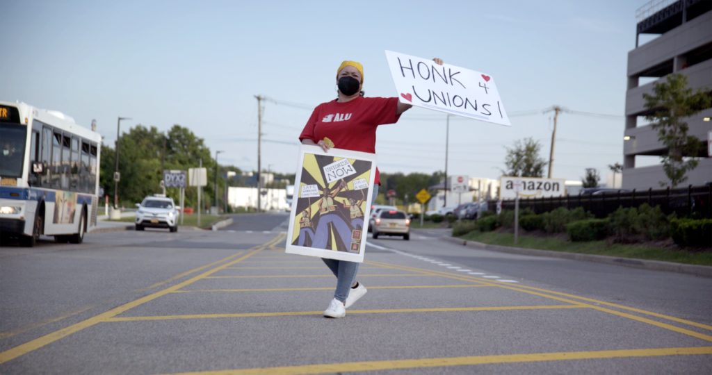 Person holding a sign that reads "Honk 4 Unions!" while standing on a road.