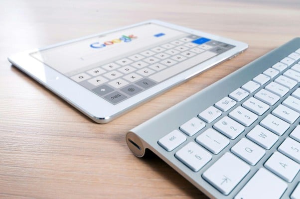 A tablet displaying a search engine interface beside a wireless keyboard on a wooden surface.