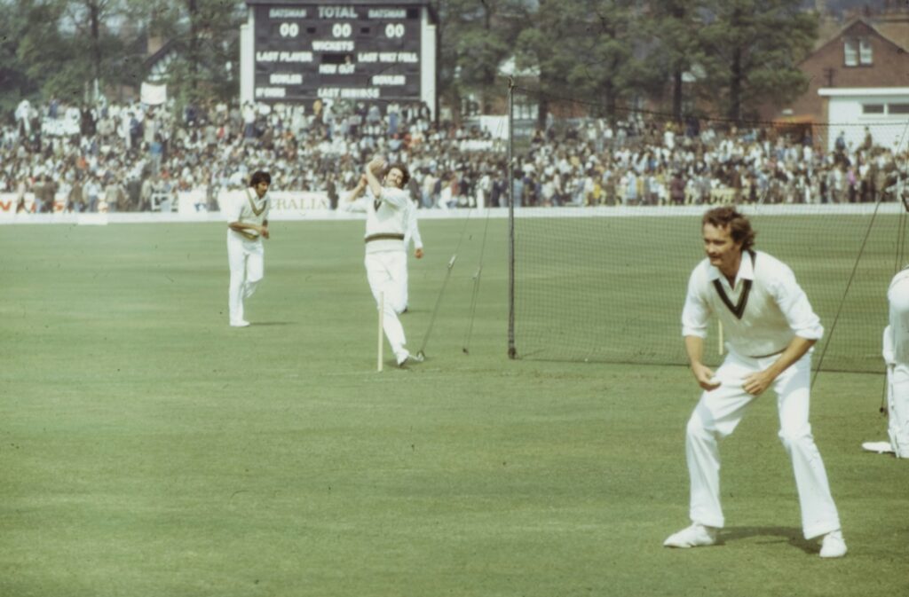 Cricket match in progress with players on the field, focused on bowling and fielding. A scoreboard and spectators are visible in the background.