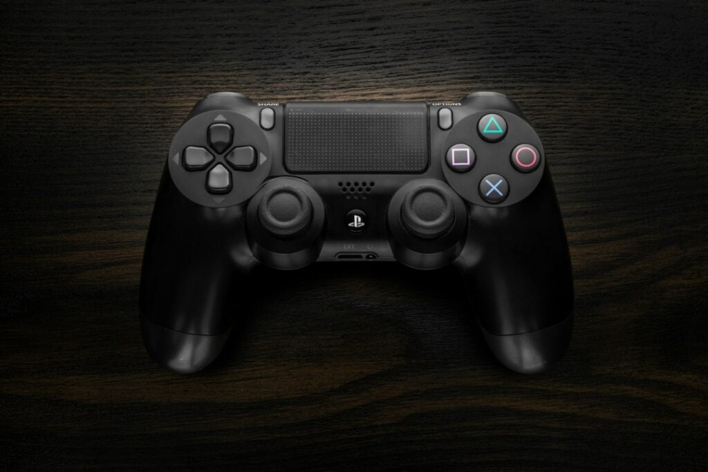 A black gaming controller with multiple buttons, including directional arrows and colored symbols, rests on a dark wooden surface.