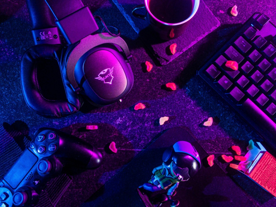 A gaming setup with a keyboard, headset, and controller on a desk. A figurine and scattered gummy candies adorn the surface, all lit by pink and purple lighting.