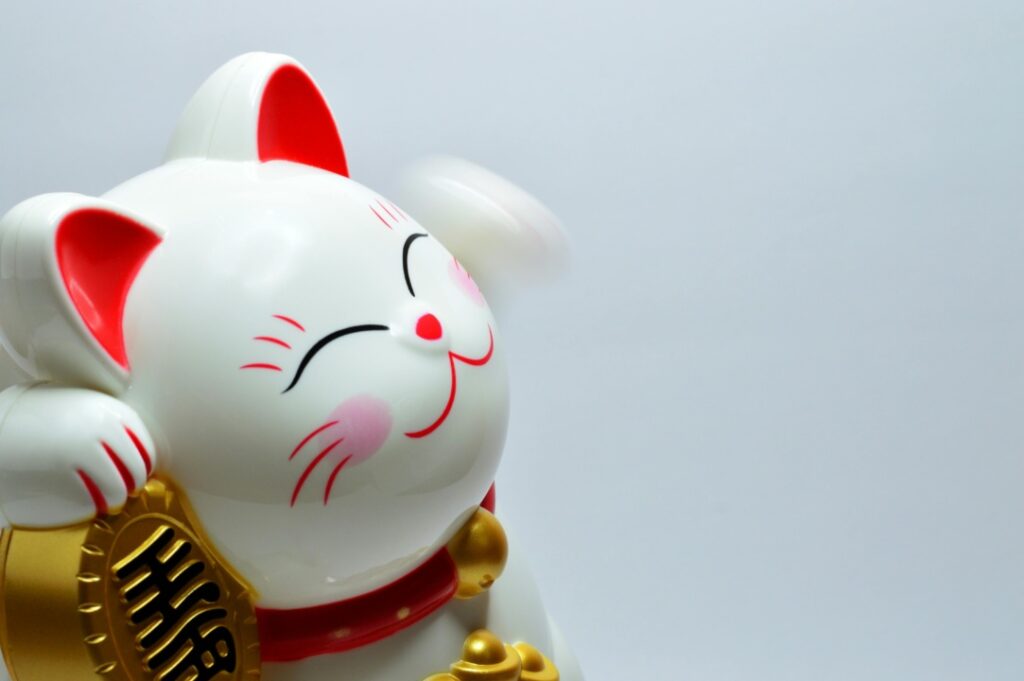 A waving white ceramic cat figurine, known as a Maneki-neko, with red ears, a red collar, and holding a gold coin.