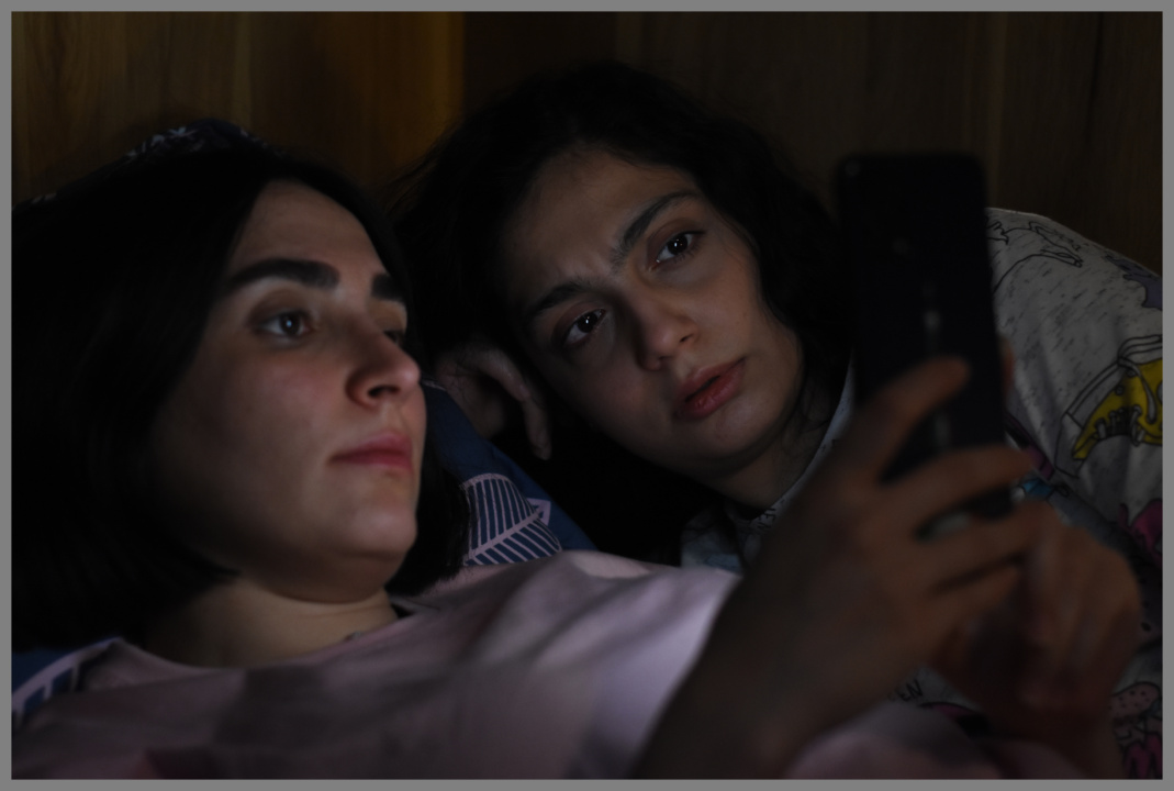 Two women are lying in bed, intently looking at a smartphone screen. One woman holds the phone while the other rests her head on her hand.
