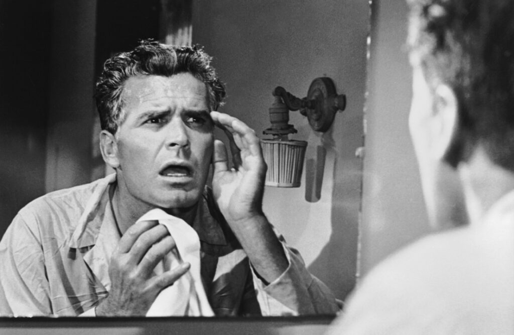 A man examines his face in the mirror, looking concerned, while holding a towel.