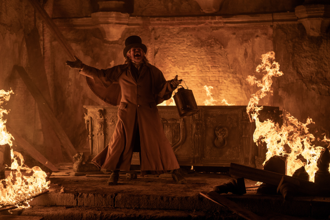 Person in a long coat and top hat stands in a dimly lit, fiery room, holding a bucket and spreading their arms. Flames surround them, casting light on the stone walls and debris.