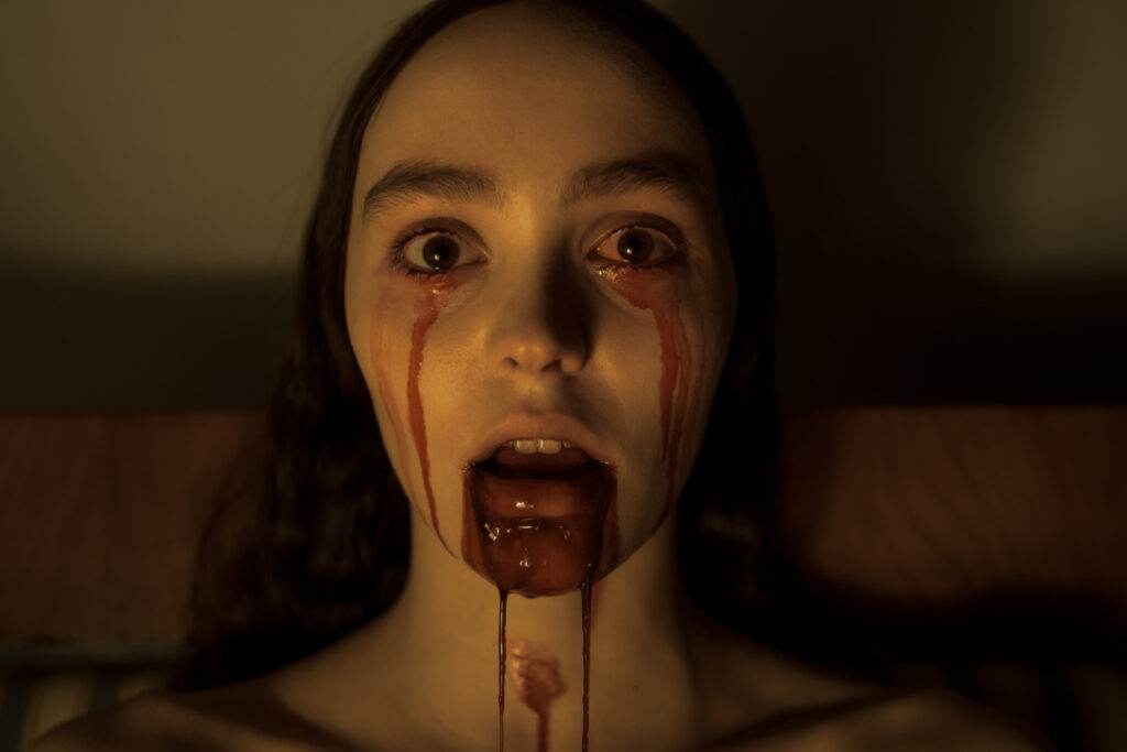 A person with long hair and an open mouth has blood streaming from their eyes and mouth, with a dimly lit background.