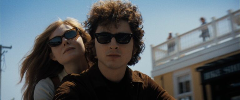 A man and woman wearing sunglasses are riding a motorcycle. The sky is clear, and there is a building with people on the balcony in the background.