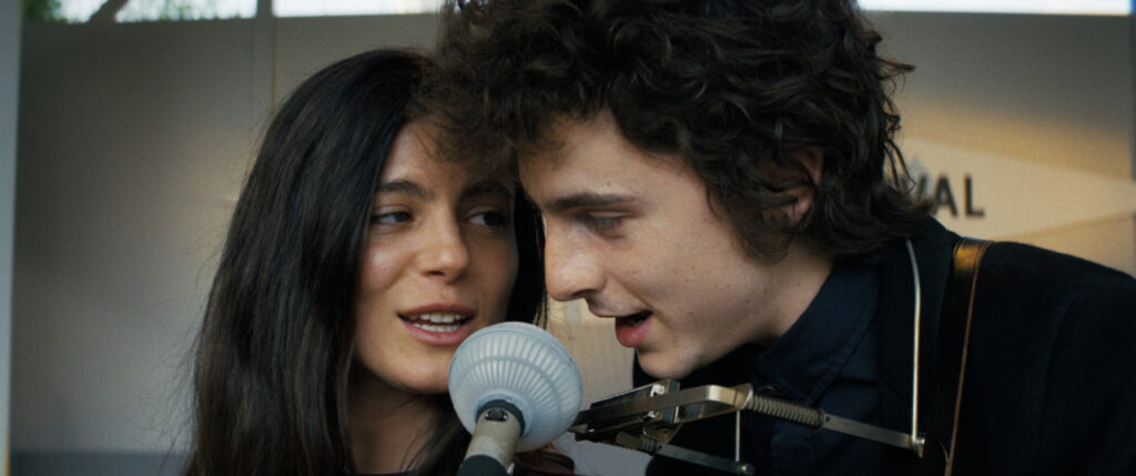 Two people sing into a vintage microphone. The person on the right has curly hair and wears a harmonica holder.