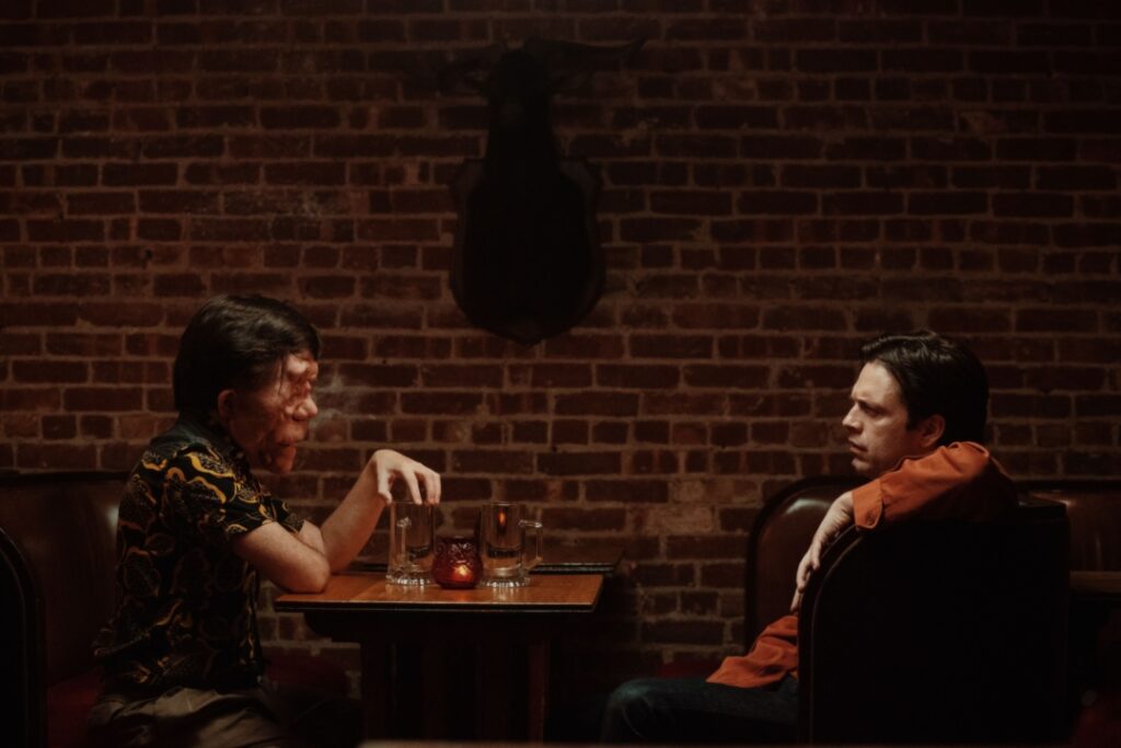 Two people seated at a wooden table in a dimly lit brick-walled room, engaged in conversation. Glasses and a small candle are on the table.