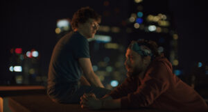 Two people sit on a rooftop ledge at night with blurred city lights in the background.