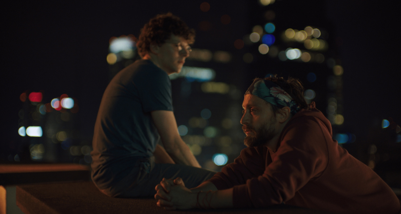 Two people sit on a rooftop ledge at night with blurred city lights in the background.