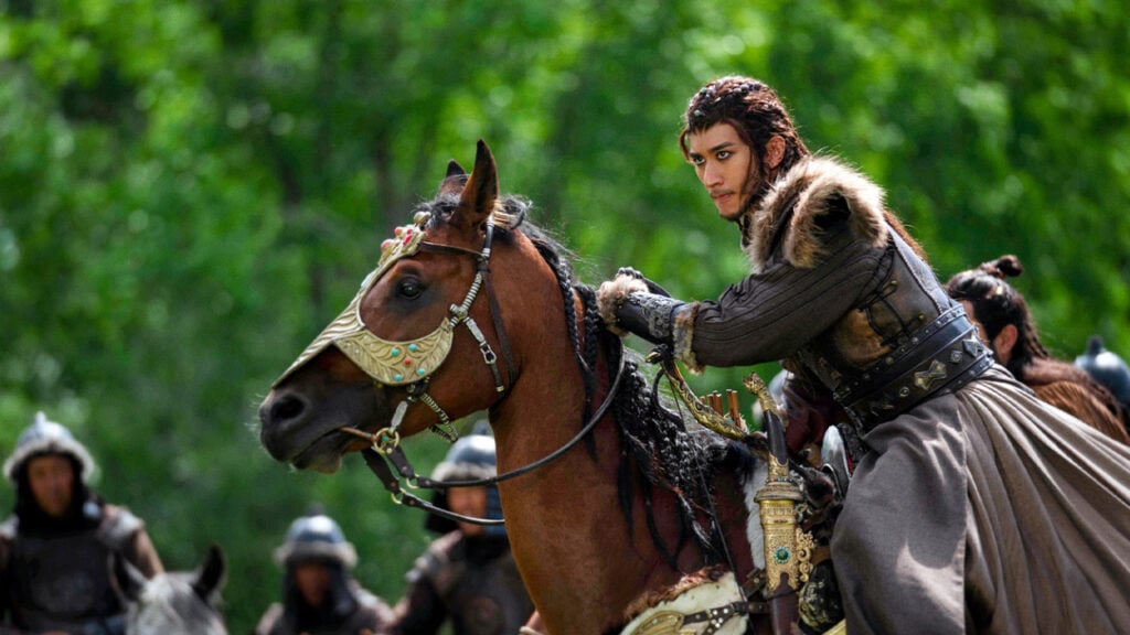 A person in historical attire rides a decorated horse; others on horseback are visible in the background against a green forest.