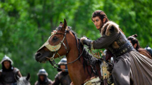 A person in historical attire rides a decorated horse; others on horseback are visible in the background against a green forest.