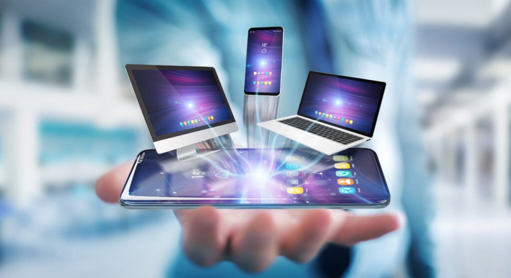 A hand holding a smartphone with holographic images of a desktop monitor, smartphone, and laptop emerging from it.