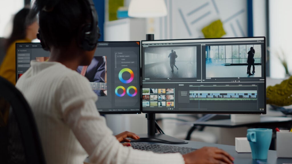 Person editing a video on dual monitors, wearing headphones, with a keyboard and coffee cup nearby.