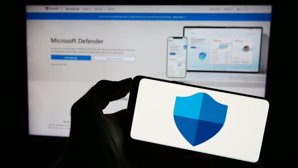 Silhouette of a hand holding a phone with a Microsoft Defender shield icon, in front of a computer screen displaying the Microsoft Defender website.